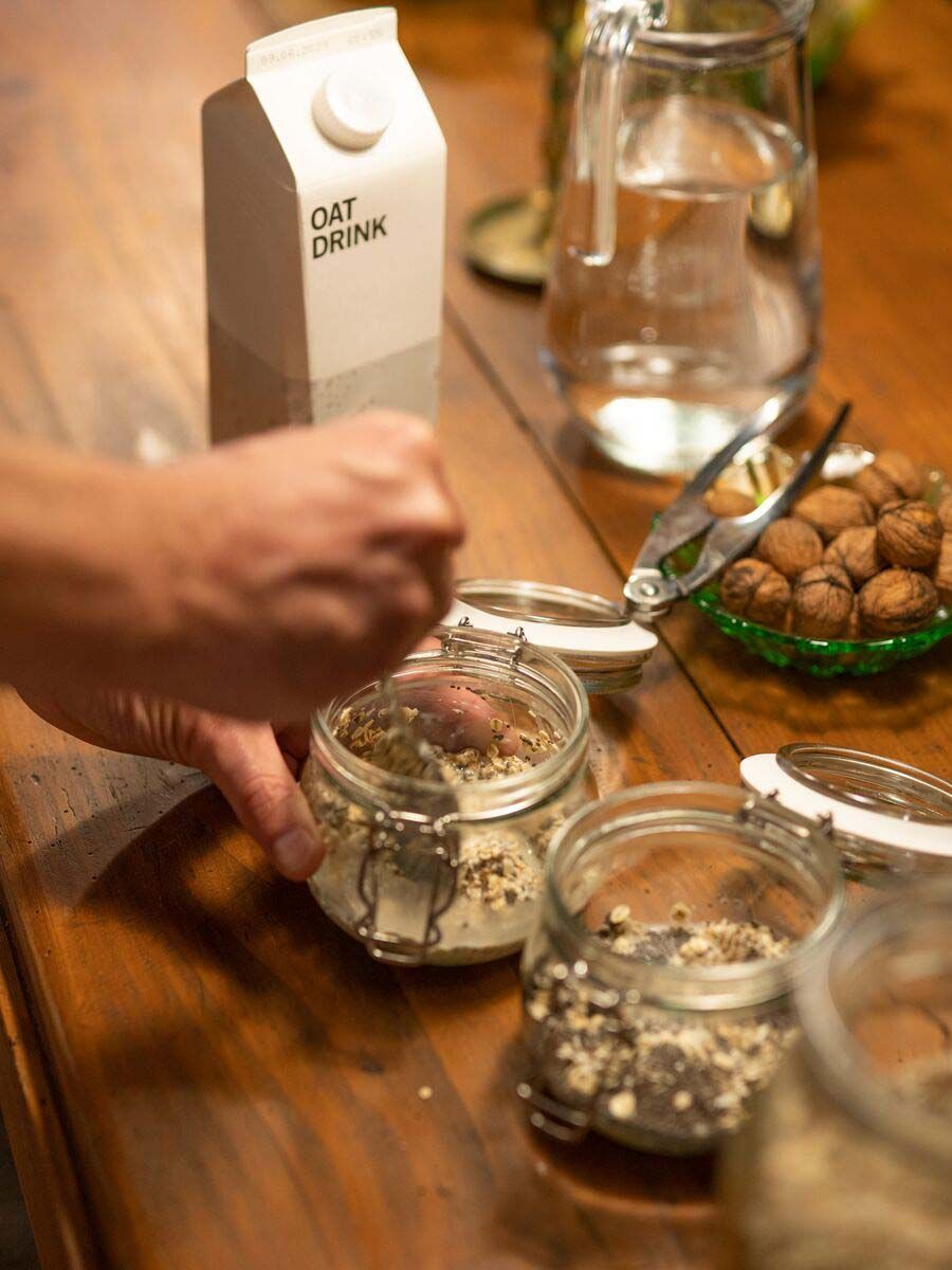 A hand spoons overnight oats ingredients into a jar. The ingredients are collected together on a wooden table.