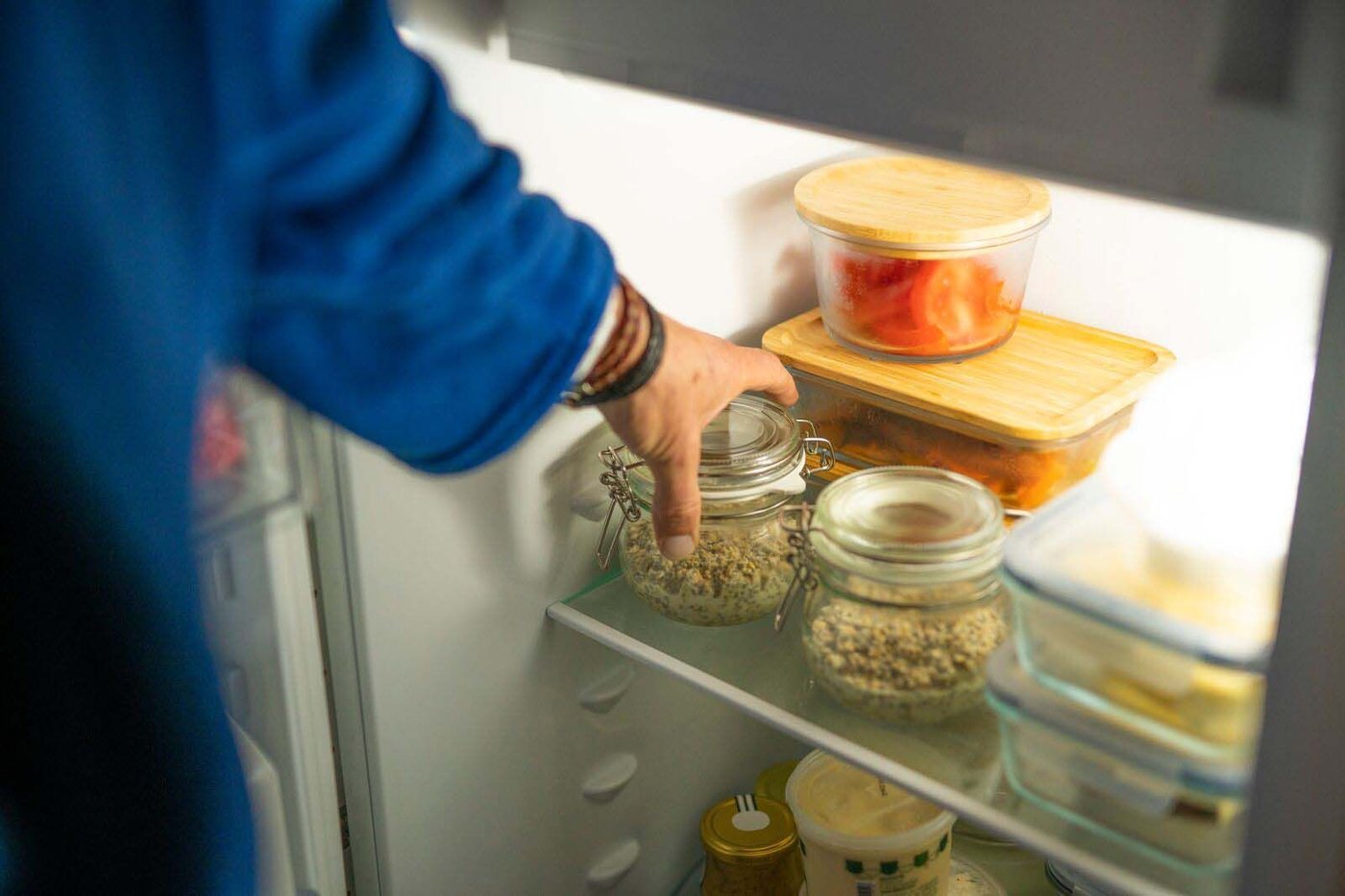 A hand reaches into the fridge. It is holding a jar filled with overnight oats ingredients.