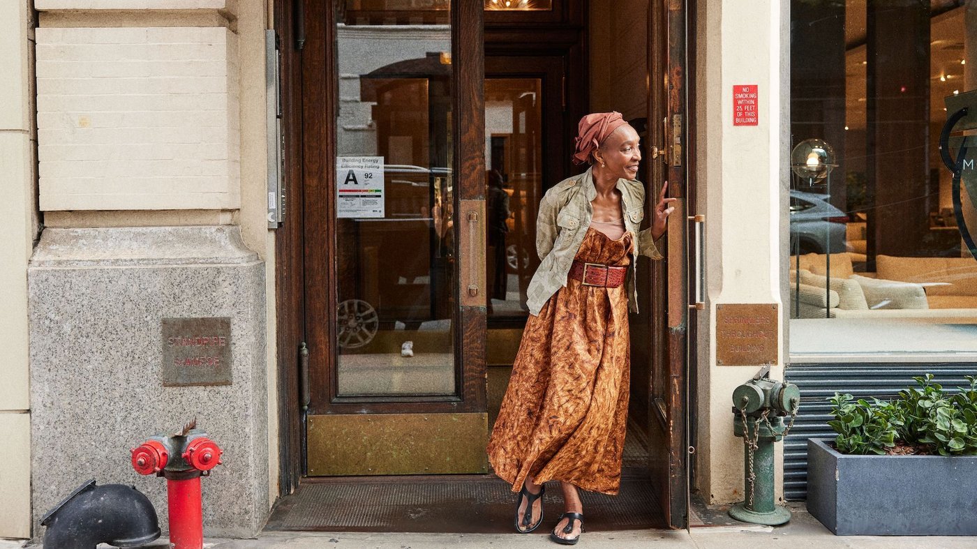 A woman in a long skirt and head scarf stands in the open doorway of a building in the city.