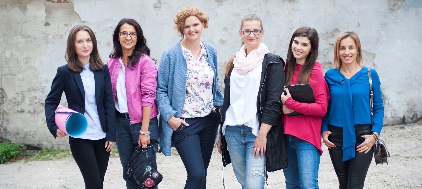 A group of women stand looking and smiling at the camera. They stand outside. One is holding paper, while another is holding a computer.