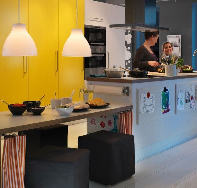 A kitchen/dining area with yellow doors and pendant lamps. Two people speak in the kitchen.