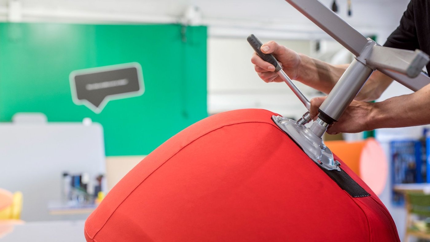 The hands of a person holding a wrench while working on an upside down swivel chair that is upholstered in orange fabric.