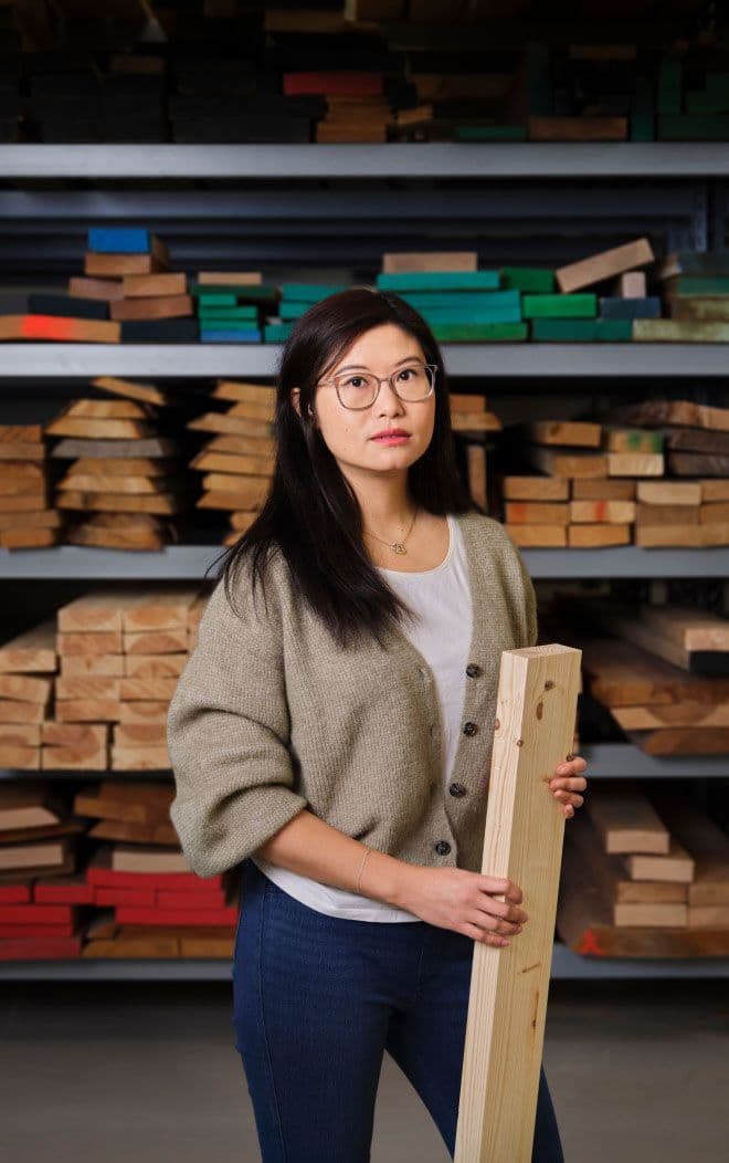 A woman stands in front of shelves stacked with wood. She is holding a piece of wood.
