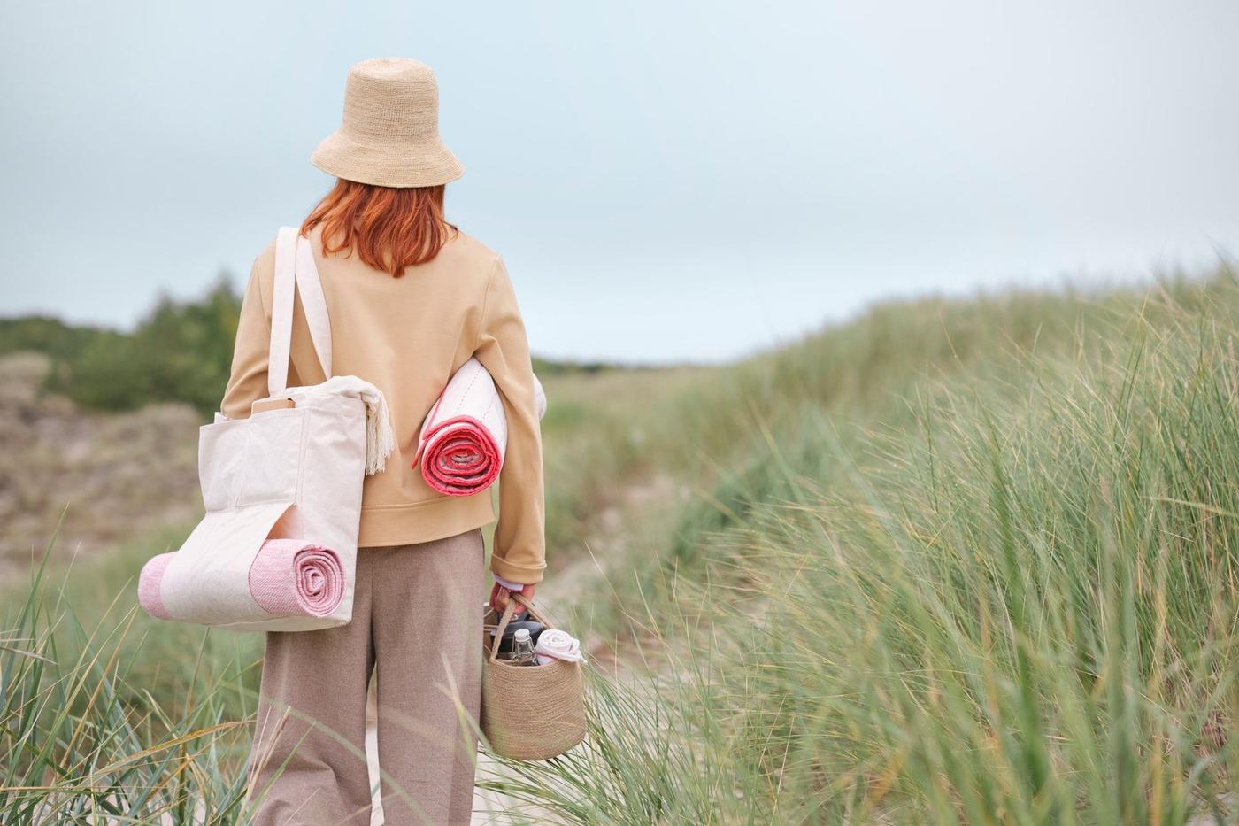 The back of a woman, dressed in a hat, carrying a bag on her left shoulder, walking away on the beach.