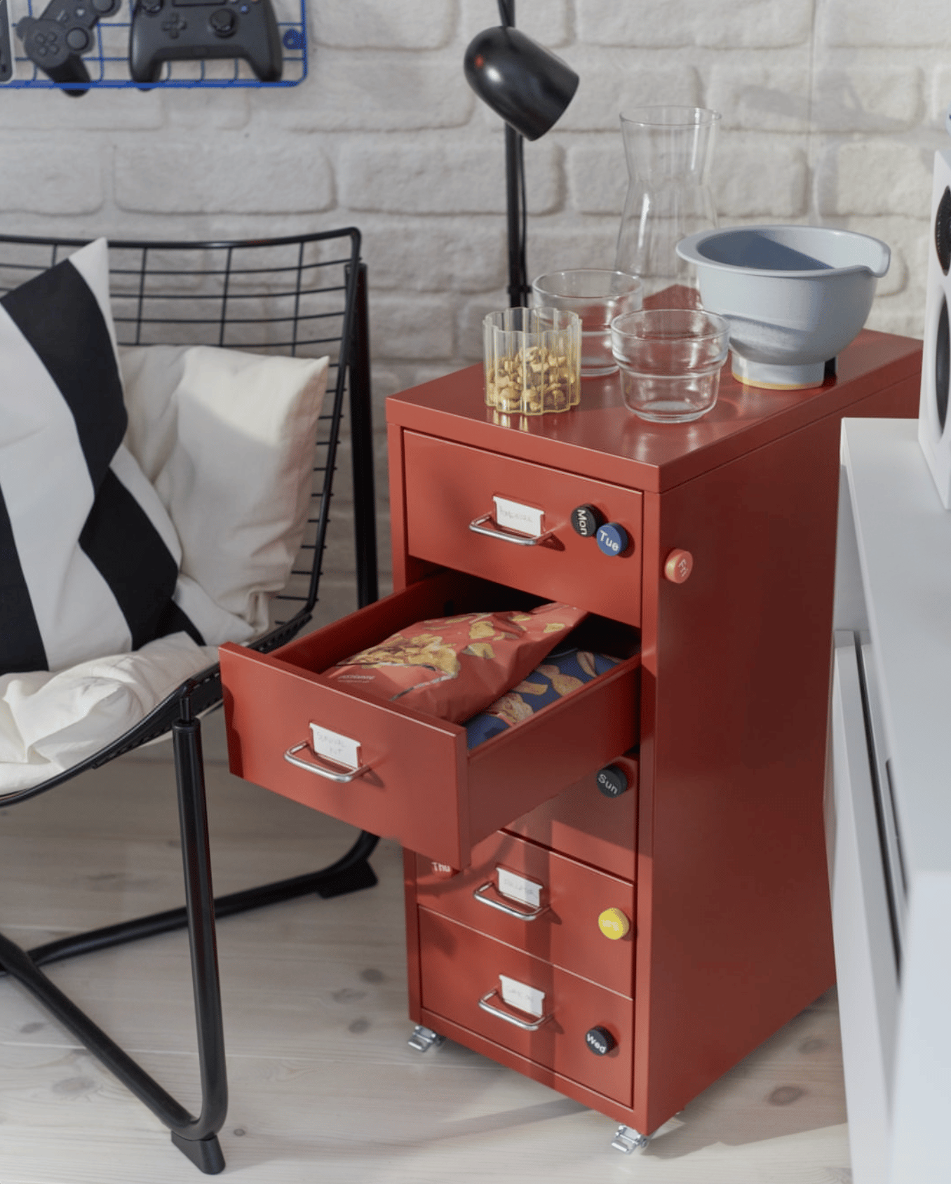 A red HELMER drawer unit on castors with an open drawer showing bags of chips and bowls and glasses sit on top with other snacks.