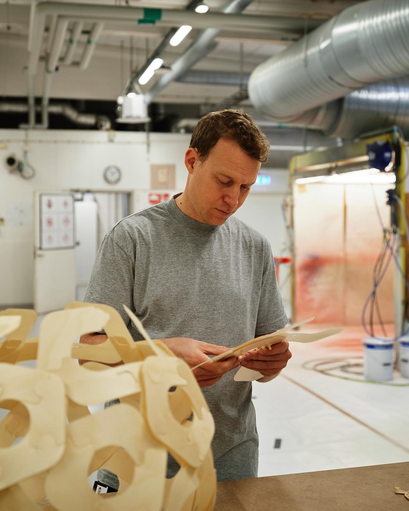 Product designer David Wahl putting together pieces of the ÖDLEBLAD lamp shade, standing beside a built ÖDLEBLAD shade, in an industrial workshop space.
