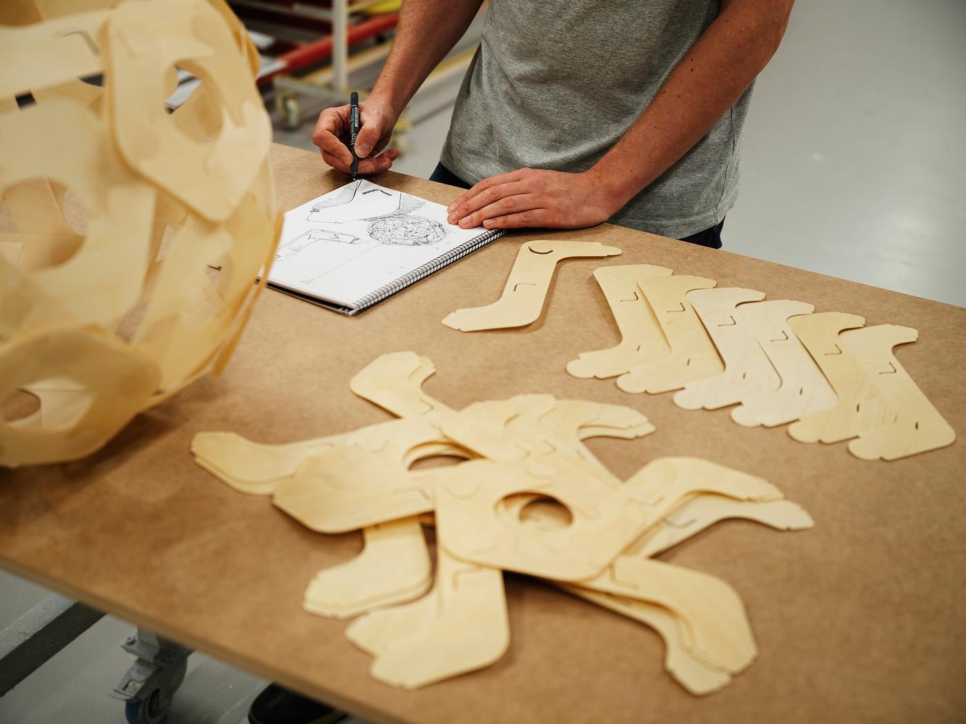 Product designer David Wahl sketching ideas for the ÖDLEBLAD lamp shade, beside one built shade and the parts of another shade, on a wood tabletop.