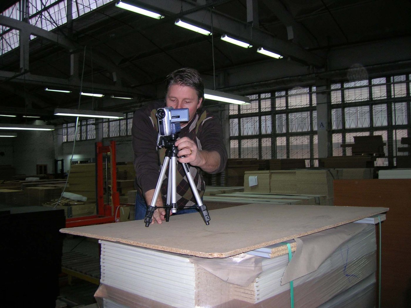 Roger looks through a video camera, set up on a tripod. The camera is on top of a plywood surface, in an industrial workspace.