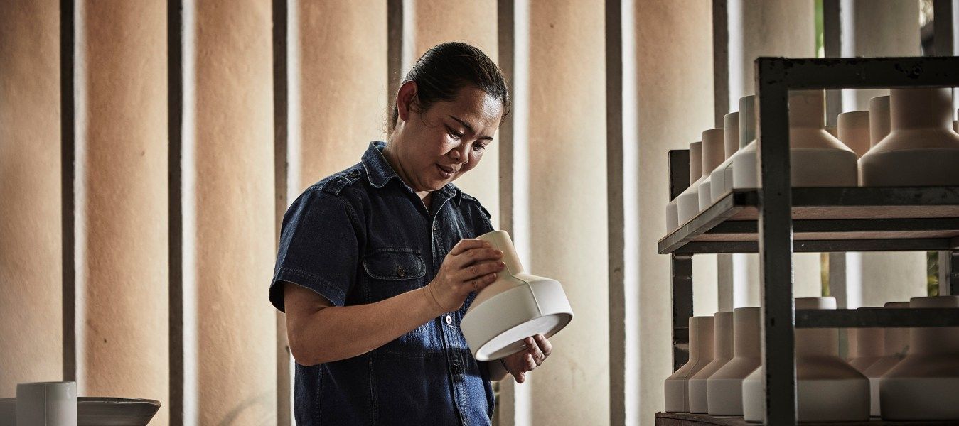 A social entrepreneur in a jeans dress is inspecting a ceramic vase next to shelves filled with vases in the same shape.