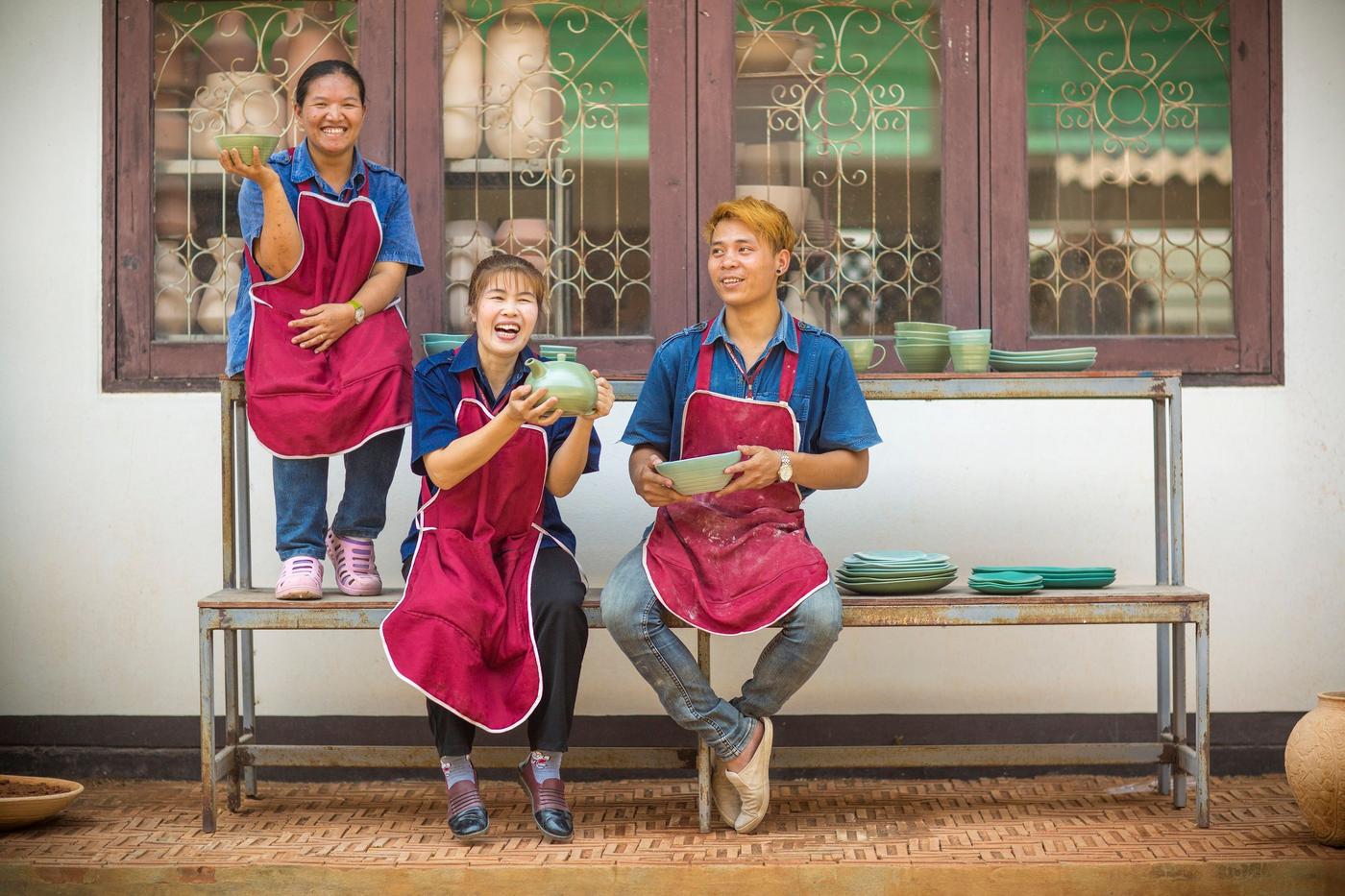 Three women social entrepreneurs with blue shirts and red aprons are sitting on a bench showing their art.