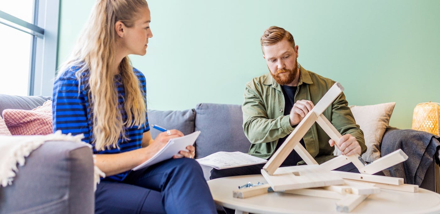 Two people looking closely at the construction of a wooden stool and taking notes while sitting in a sofa next to a window.