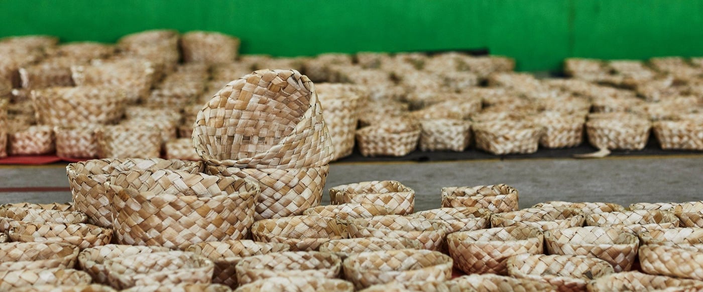 Around one hundred handcrafted straw baskets in natural shades of beige standing on the ground in front of a green wall.