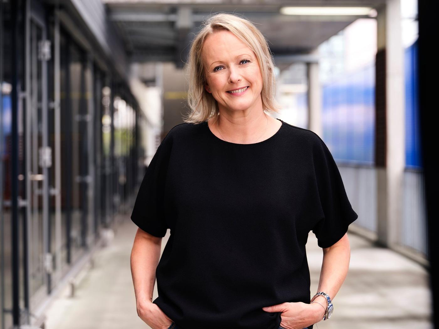 Portrait of Susanne Waidzunas, who is wearing a black shirt and smiling at the camera with her hands in her pockets.