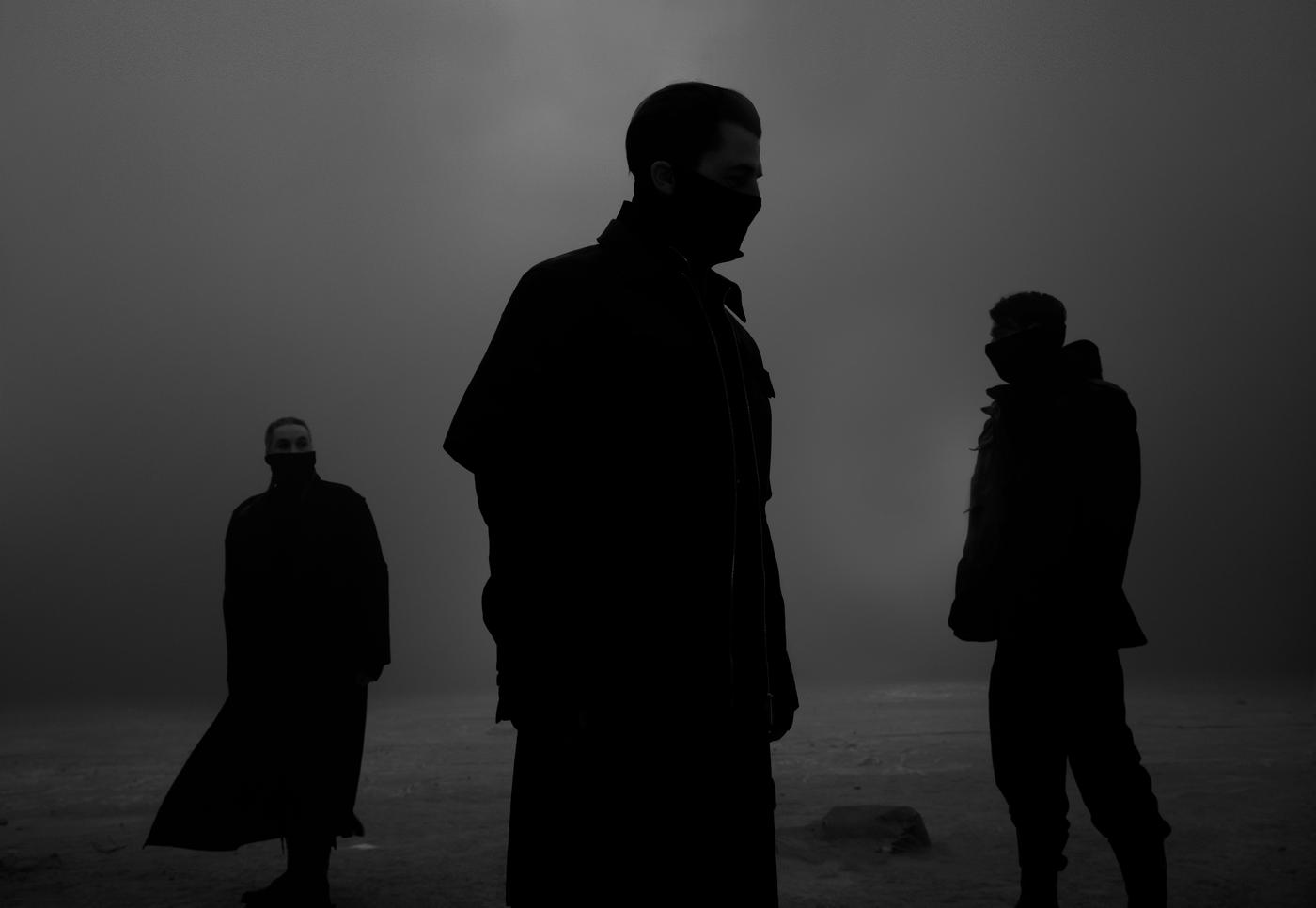 In a dimly lit grey space, the silhouettes of three people standing.