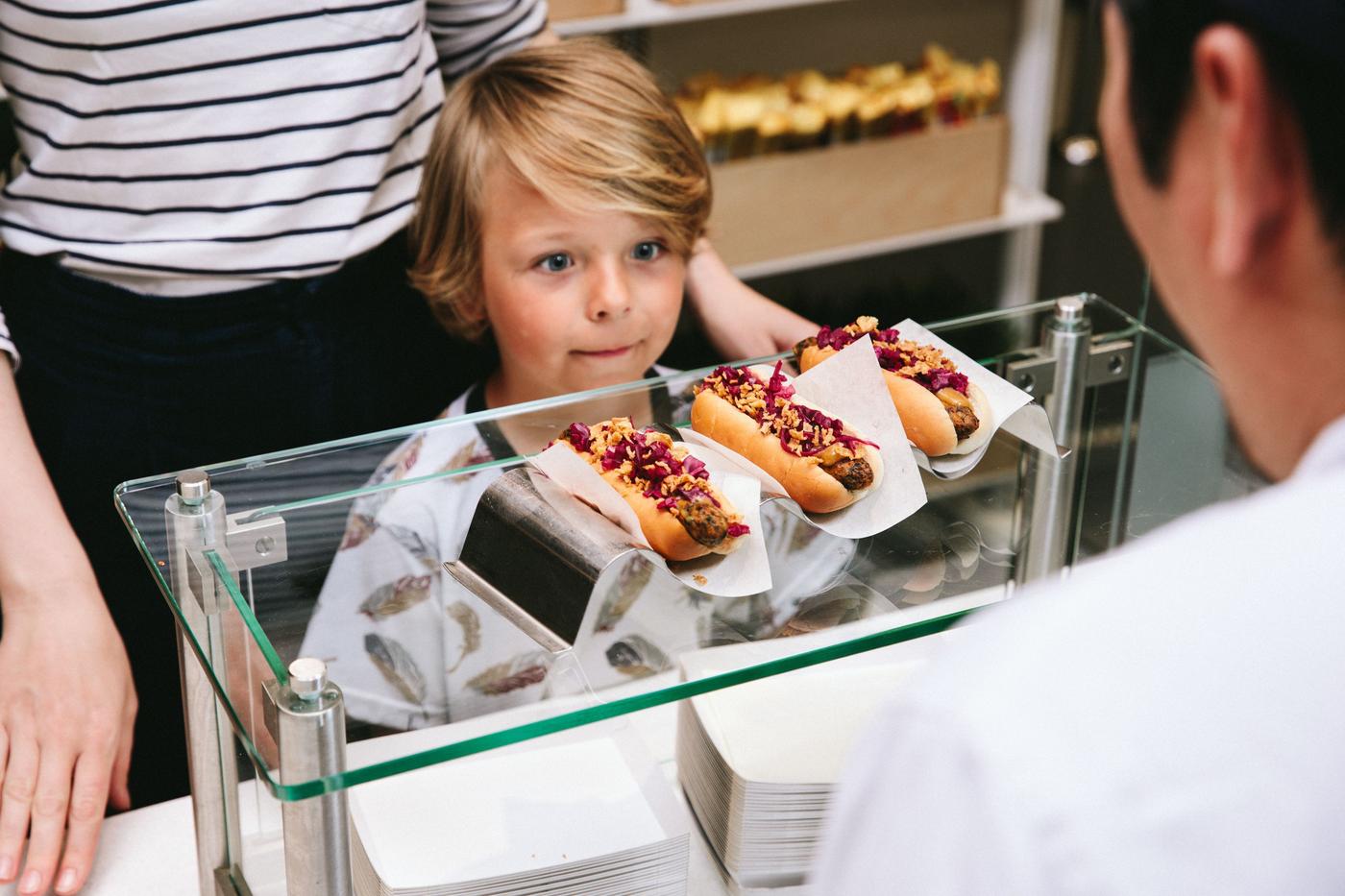 A little boy with a hungry and happy expression is looking at three veggie hot dogs being prepared by a Bistro co-worker.
