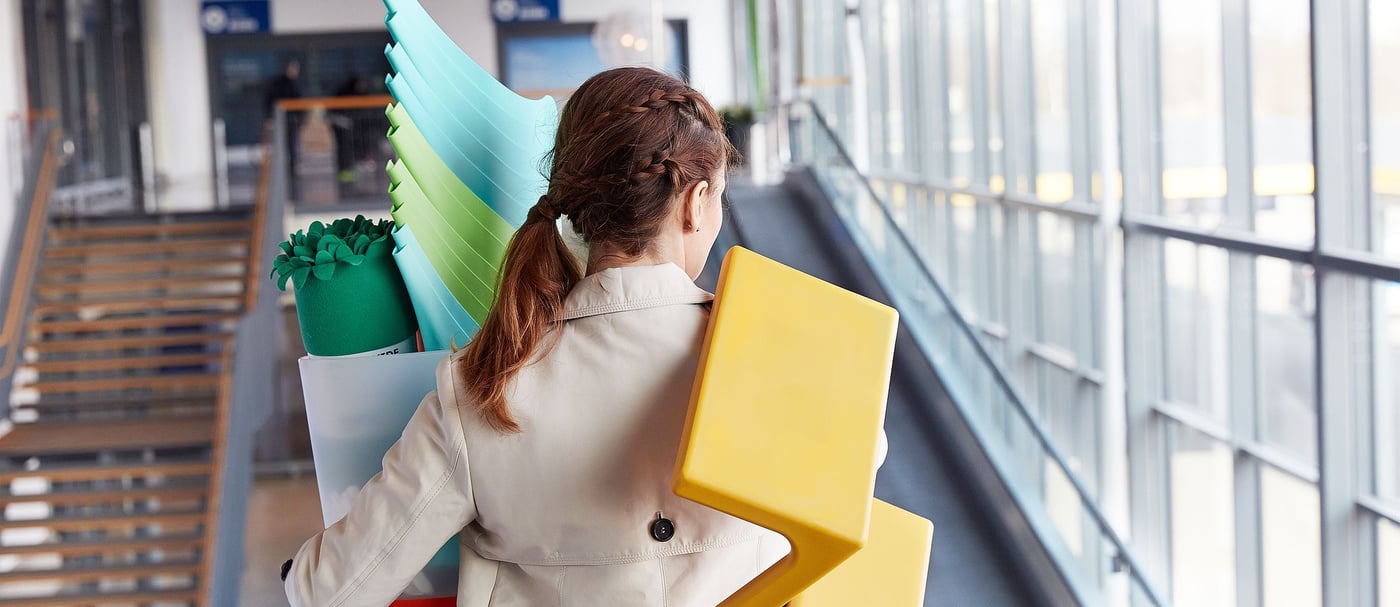 A woman in a beige trench coat carrying a pile of colourful watering cans. A yellow stool is hanging over her shoulder.