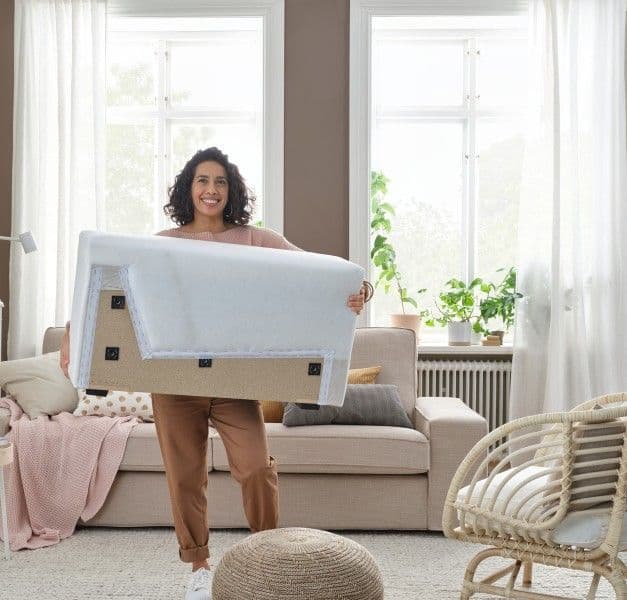 A woman holding a sofa part without cover in a white-beige living room with a sofa, armchair, throw, table, and plants.