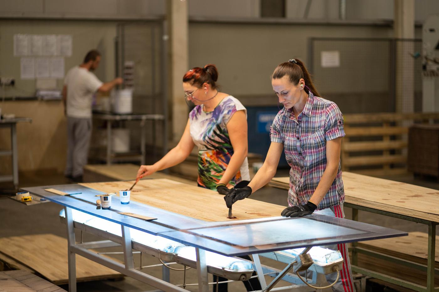 Two women in a factory working treating a large sheet of pine.