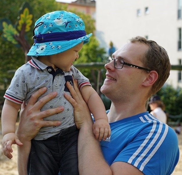 A man wearing glasses and a blue shirt holds and smiles at a baby wearing a blue bucket hat.