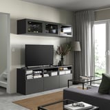 Moderan dnevni boravak s BESTÅ crnim TV ormarićem i zidnim ormarićima. TV ravnog ekrana, uredno raspoređene knjige i sivi tepih. Veliki prozor sa sivim zavjesama.