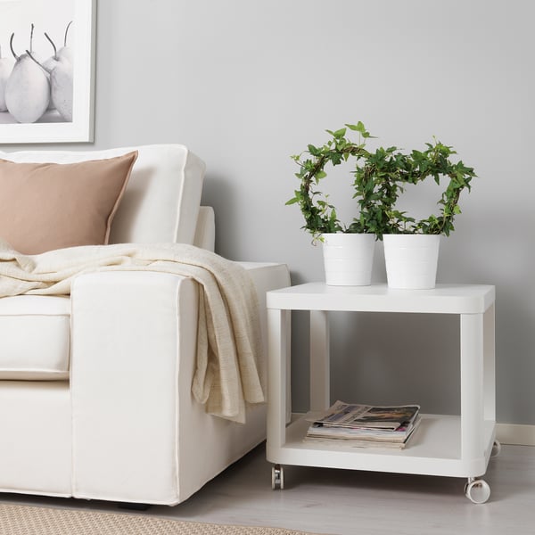 Udoban bijeli kauč s bež jastucima i dekom, pored malog bijelog pomoćnog stolića s dvije biljke u saksiji i hrpom časopisa.