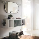 Bijeli regal KALLAX s mrežastim ladicama, ispod velikog ovalnog ogledala, u blizini svijetlih vrata.