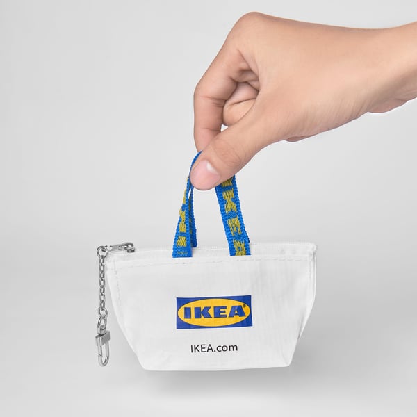 U ruci se nalazi mala bijela IKEA torbica s plavim patentnim zatvaračem i pričvršćenom metalnom karikom lanca, idealna za odlaganje novčića i sitnica.