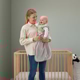 Mama se smiješi s bebom u sivoj vreći za spavanje, dva krevetića u zelenoj dječjoj sobi.