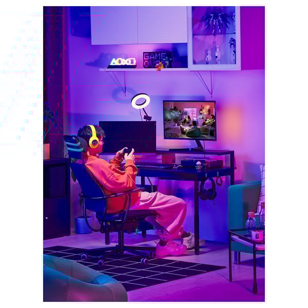 Gamer sjedi u sobi s neonskim osvjetljenjem, nosi slušalice, igra videoigre na računalu, sa stolicom LÖPARBANA, stolom i prstenastim svjetlom.