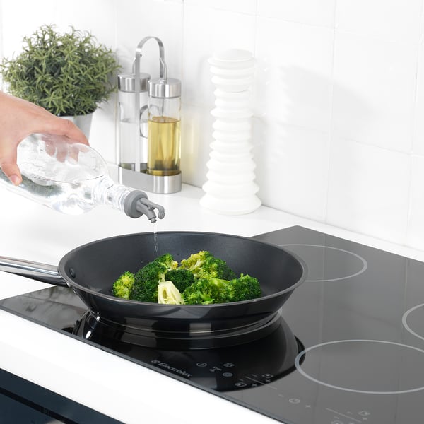Ruka ulijeva vodu iz boce u tavu s brokulom na ploči za kuhanje pomoću točila LÖSTAGBAR.
