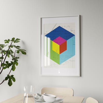 Raznobojna geometrijska umjetnost u bijelom okviru iznad blagovaonskog stola s bijelim posuđem i biljkom.