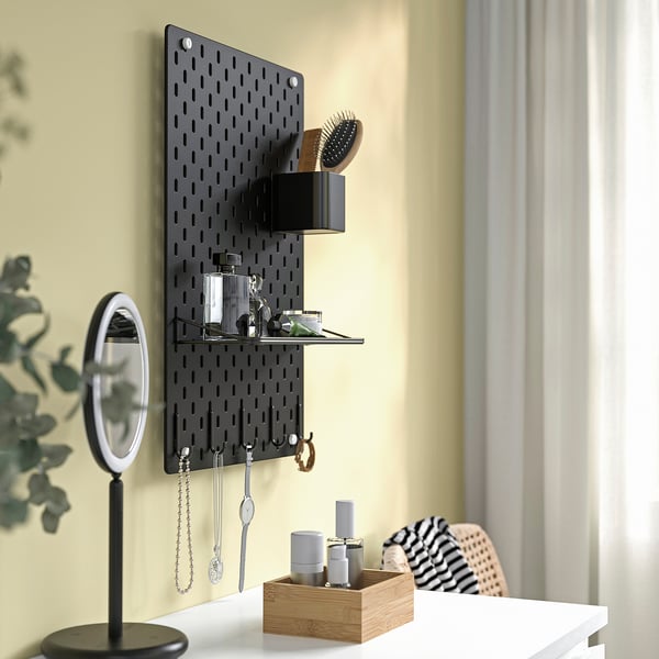 Crna SKÅDIS pegboard s kukama, policama i pretincima u kojima se nalaze kozmetički predmeti, nakit i biljke na žutom zidu.