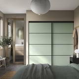 Moderna spavaća soba s SKYTTA zelenim ormarom od matiranog stakla s kliznim vratima. Krevet svijetle boje i bež zidovi poboljšavaju minimalistički dizajn.