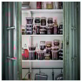 Polica s raznim staklenkama, mnoge ispunjene tamnim sadržajem nalik konzervi, u ugodnoj, rustikalnoj sobi s tirkizno zelenim vratima.