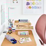 Šareni ormarić za stol od pluta s raznim uredskim predmetima poput olovaka, naljepnica i bilježnica, na bijelom stolu s crvenom stolicom.