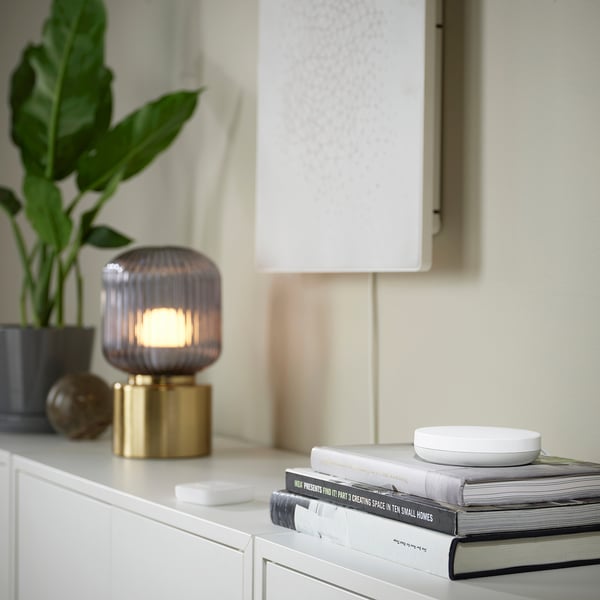 Mali bijeli daljinski upravljač za zvuk SYMFONISK na hrpi knjiga, pored elegantne svjetiljke i biljke.