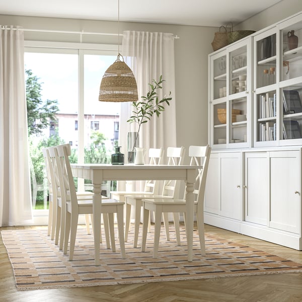 Svijetla blagovaonica s bijelim namještajem, toplim tepihom, tkanim svjetlom, biljkom i velikim prozorom.