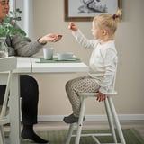 A lány átadja a tárgyat a nőnek, miközben a gyerekszéken ül.