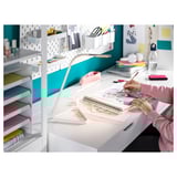 Egy személy rajzol egy fehér ALEX íróasztalnál, beépített fiókokkal és polcokkal. Az íróasztal elegáns, minimalista dizájnnal rendelkezik, mögötte egy kékeszöld fallal.