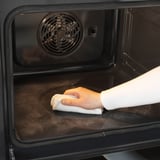 A személy ruhával tisztítja a rozsdamentes acél sütő belsejét. Öntisztító funkció és ujjlenyomat-álló felület látható.