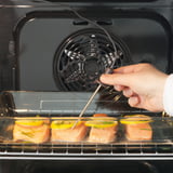 Egy kéz fogóval citrommal díszített lazacfilét helyez egy nyitott sütőbe, légkeveréses fűtéssel.