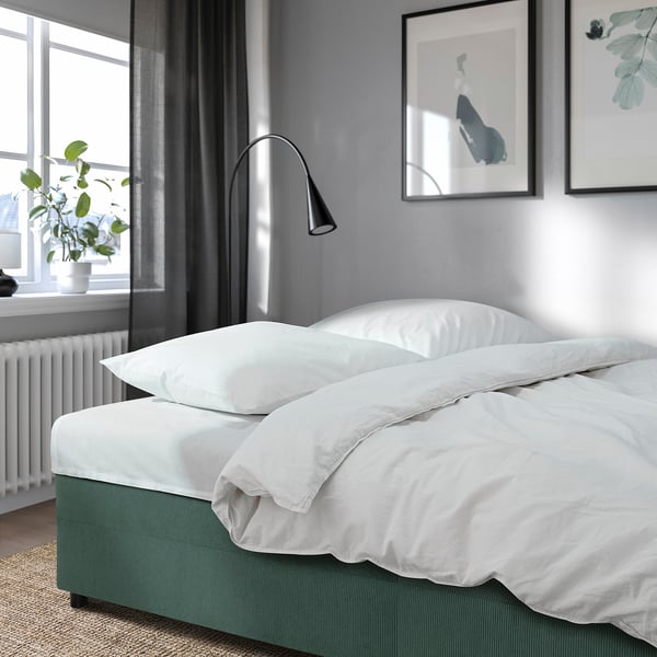 Modern minimalista hálószoba szürke falakkal, nagy ablakkal, zöld kanapéval, fehér ágyneművel és lámpával.