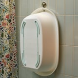 Fehér babakád puha szélekkel lóg a csempézett falon, virágfüggöny a közelben.