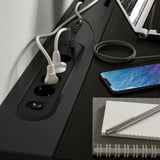 Fekete íróasztal konnektorokkal, usb-portokkal és kábelrendezővel. Telefon, notebook és tekercselt kábel a tetején.