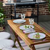 Fából készült kültéri étkezőasztal szett tányérokkal, villákkal és poharakkal, ideális nyári partikra.