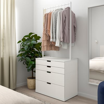 Modern hálószoba fehér IKEA NORDLI ládával. Ruhatartó rúd fent, növény mellett, ágy a közelben. Letisztult és minimalista dizájn.