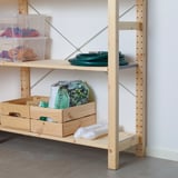 Világos fa tárolópolcok átlátszó edényekkel, amelyek színes tárgyakat tartalmaznak, beleértve a zöld tömlőt és a műanyag zacskókat az alsó polcon.