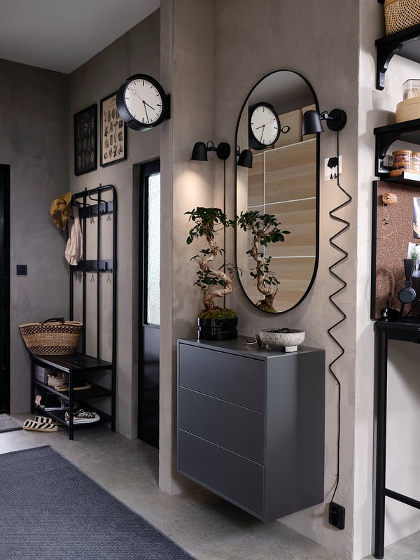 Modern bejárat szürke falakkal, SKURUP fekete állítható lámpákkal, nagy ovális tükörrel, fa ruhafogassal és szürke fiókos elemmel.