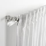 Közeli kép a fehér függönyöket tartó krómozott függönyrúd-tartóról, a soldvärm szett egy részének bemutatásával.