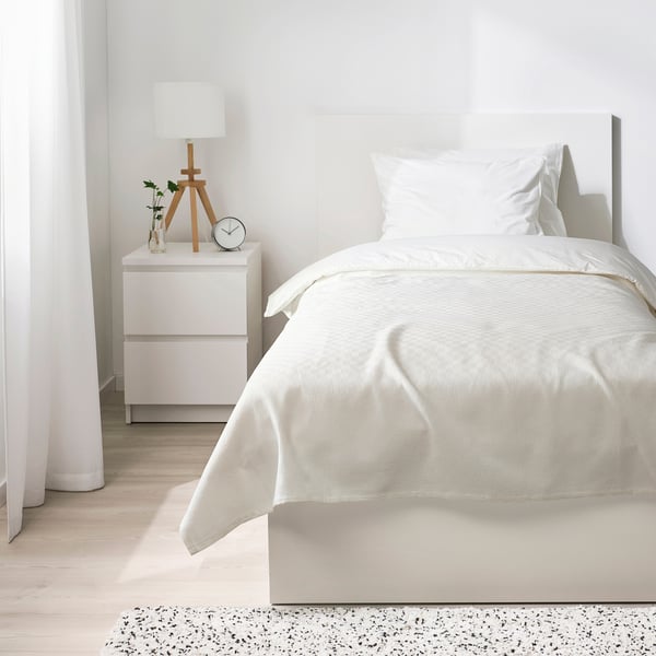 Modern hálószoba fehér STENFRÖMAL ágytakaróval, egyszemélyes ágyon, világos fa éjjeliszekrényrel, lámpával, vázával és órával. Tágas és minimalista kialakítás.